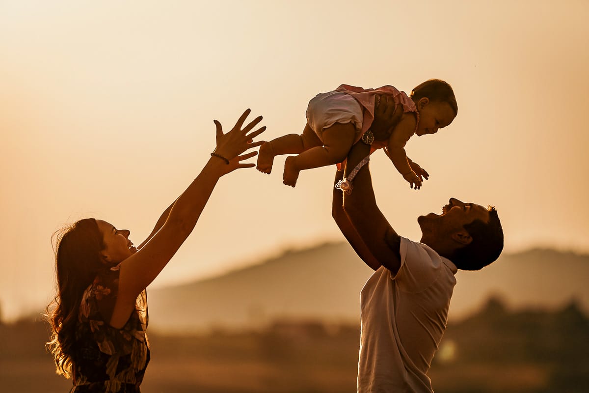 Fotografia di famiglia a Chieri. Ritratti spontanei in esterno nelle colline intorno alla città, al tramonto. Silhouette del papà che fa volare la bambina in controluce e la mamma protende le braccia.