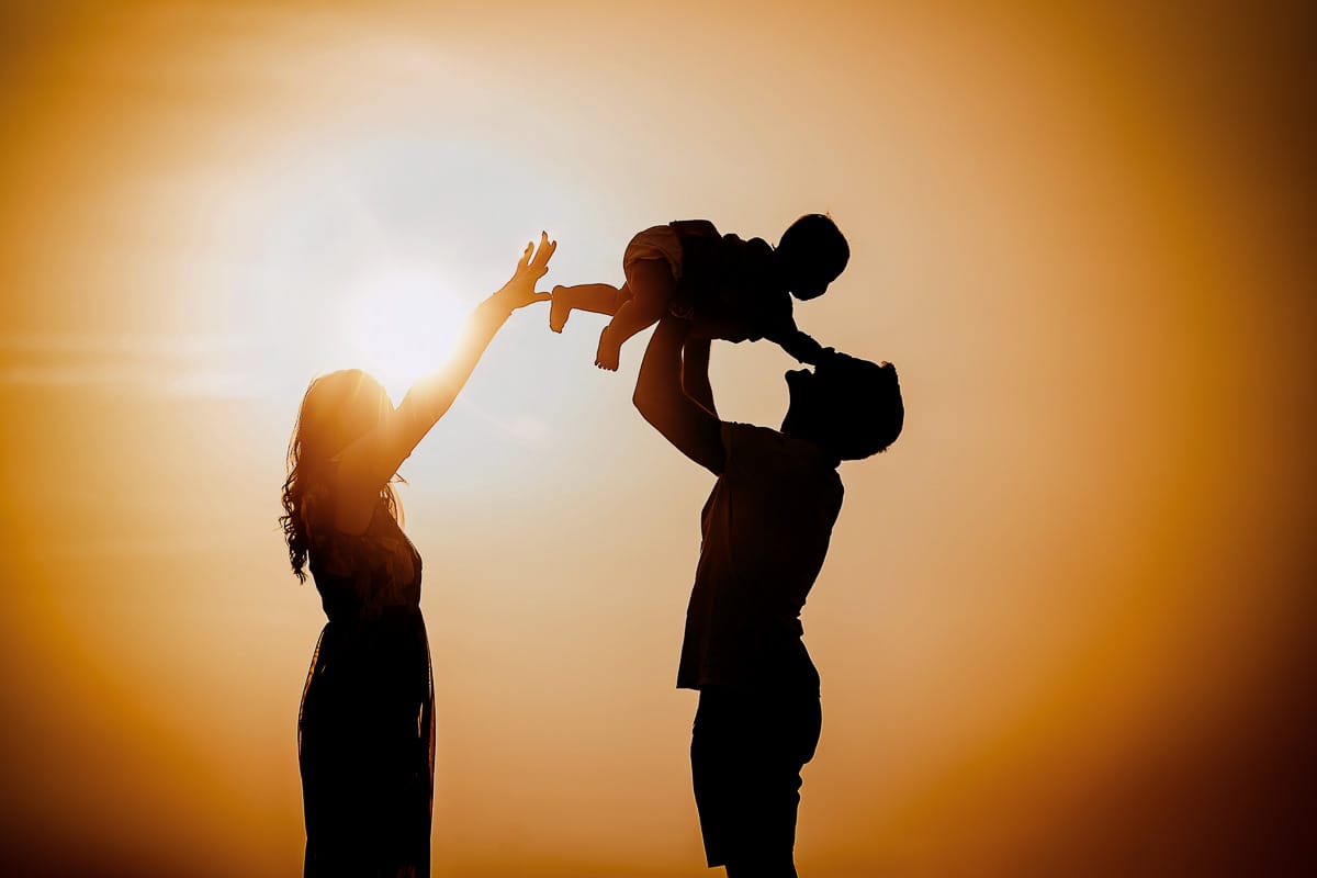 Fotografia di famiglia a Chieri. Ritratti spontanei in esterno nelle colline intorno alla città, al tramonto. Silhouette del papà che fa volare la bambina in controluce e la mamma protende le braccia.