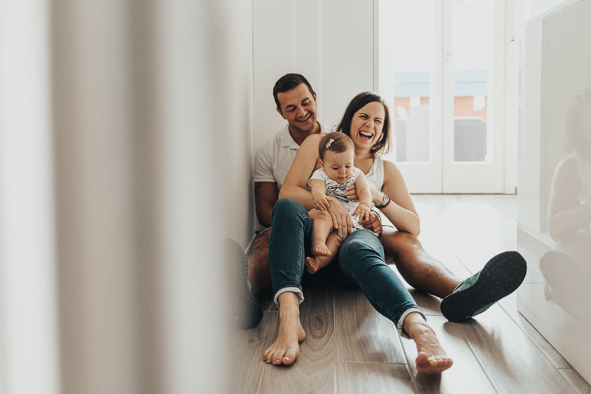 Fotografia di famiglia a Chieri. La piccola Aurora in braccio al papà con la mamma, seduti per terra in cucina, riflessi nel bancone della cucina,