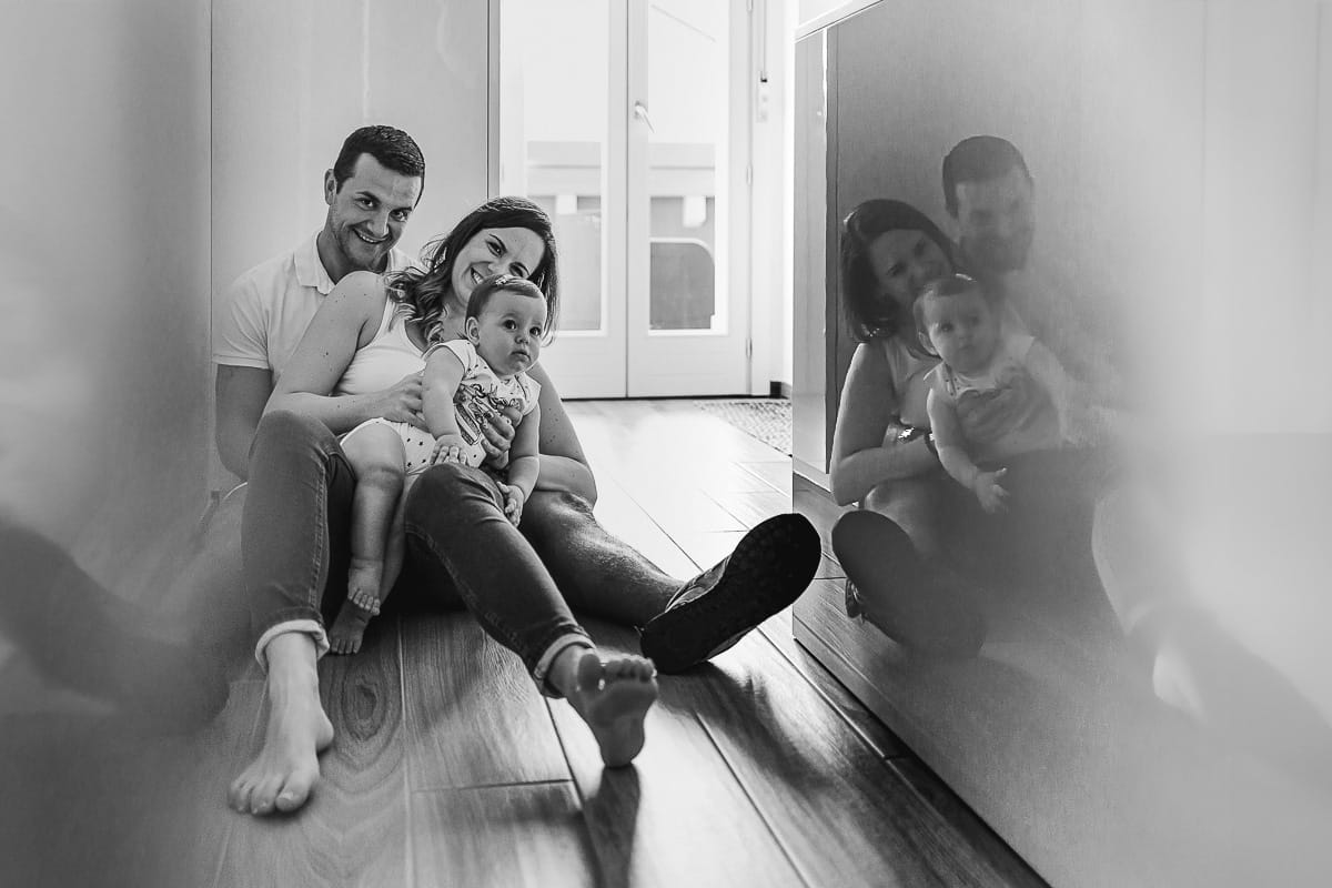 Fotografia di famiglia a Chieri. La piccola Aurora in braccio al papà con la mamma, seduti per terra in cucina, riflessi nel bancone della cucina, bianco enero