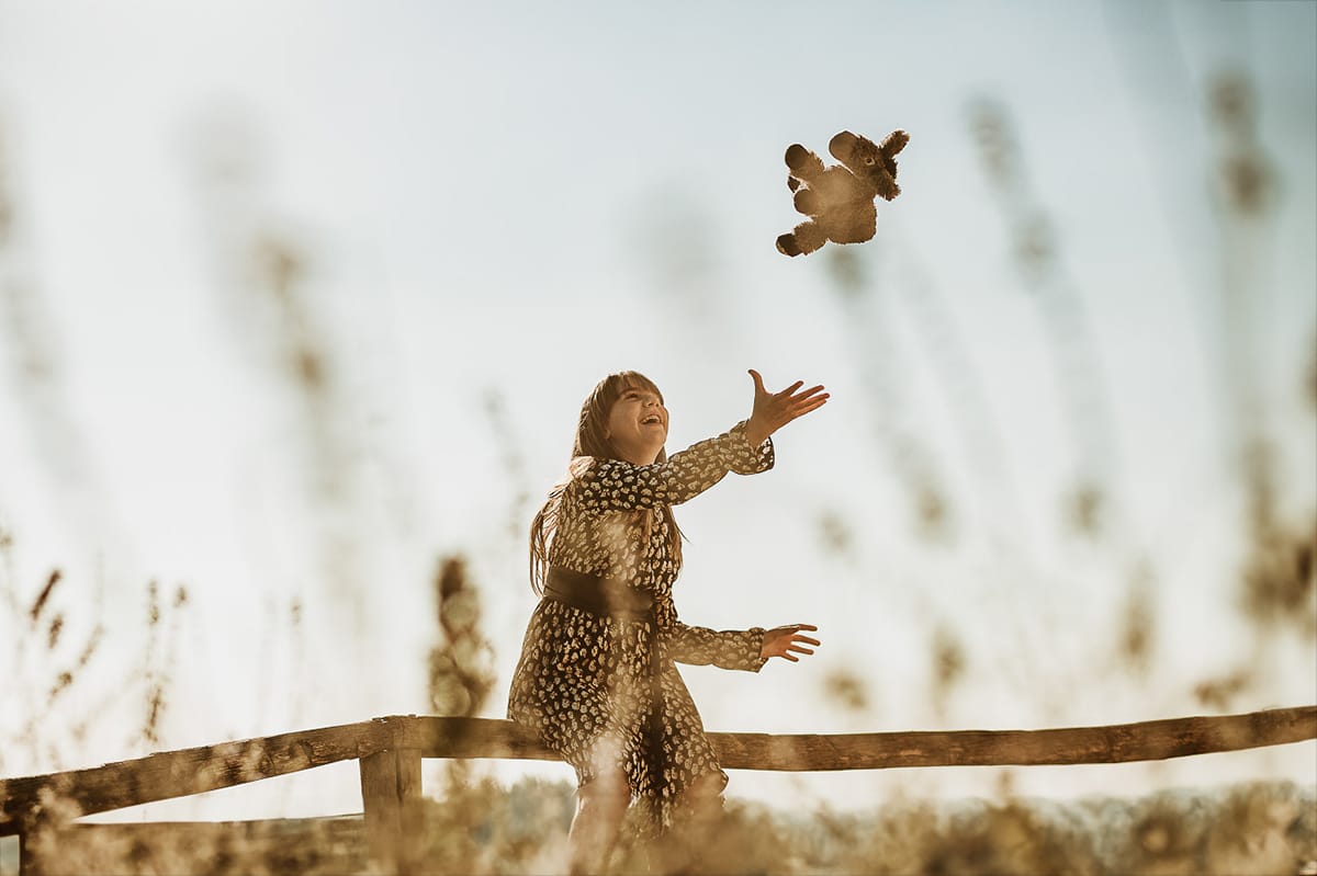 Servizio fotografico di famiglia autunnale in Monferrato. Figlia seduta su di una staccionata di legno fa volare un peluche