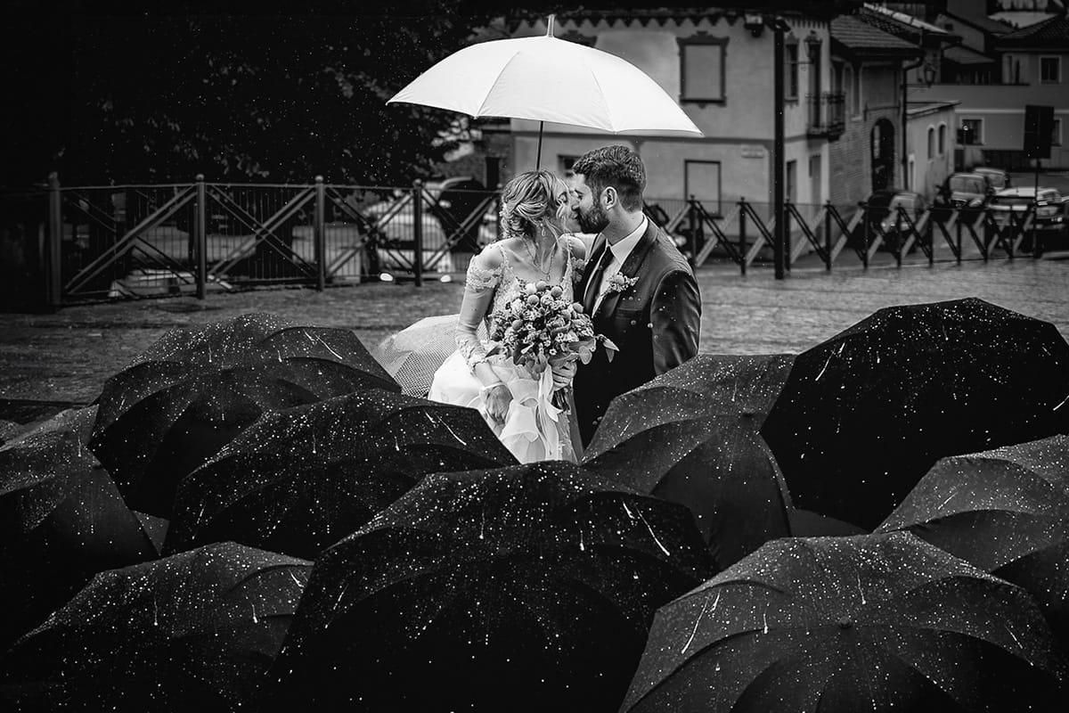 Matrimonio sotto la pioggia a Cambiano. Ritratto degli sposi sotto l'ombrello bianco. Gli invitati accucciati sotto gli ombrelli scuri