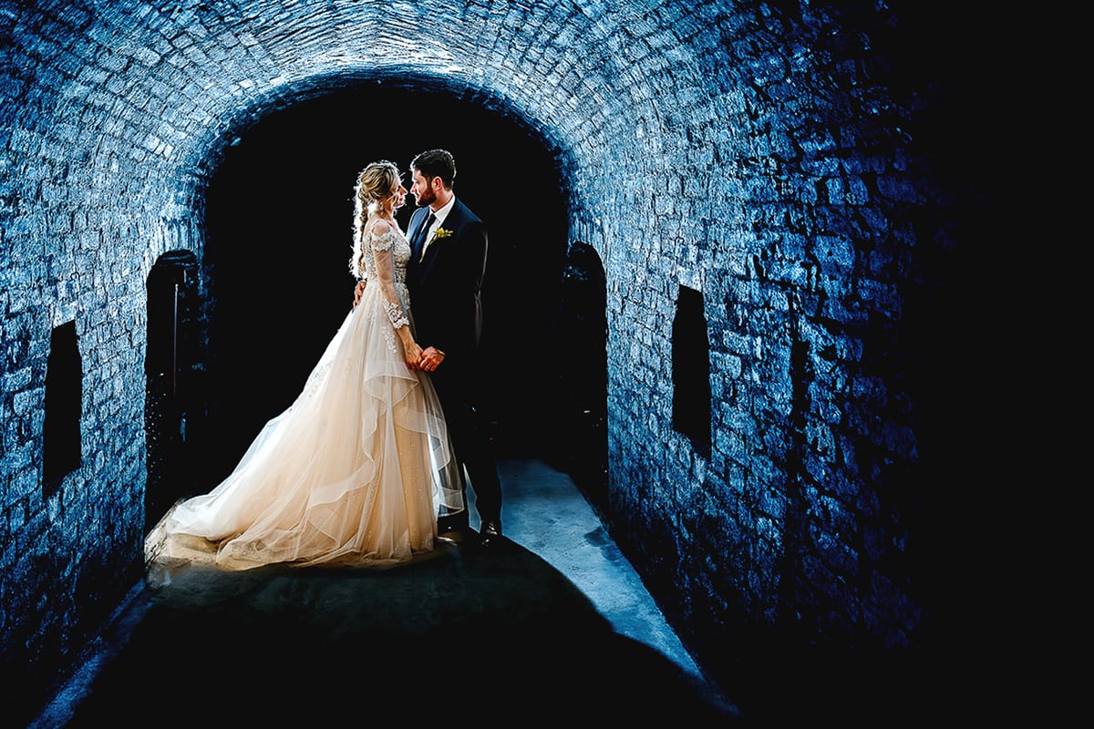 Matrimonio sotto la pioggia a Tenuta Tamburnin di Castelnuovo Don Bosco. Ritratto degli sposi nella cantina dell'azienda vitivinicola Flash esterno dietro gli sposi con gelatina blu