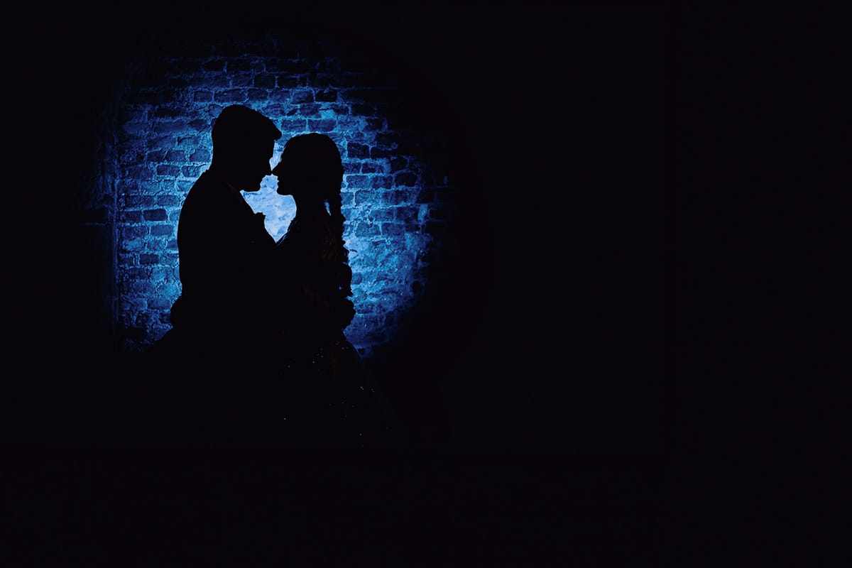 Matrimonio sotto la pioggia a Tenuta Tamburnin di Castelnuovo Don Bosco. Ritratto degli sposi nella cantina dell'azienda vitivinicola Flash esterno dietro gli sposi con gelatina blu che crea una silhouette sul muro