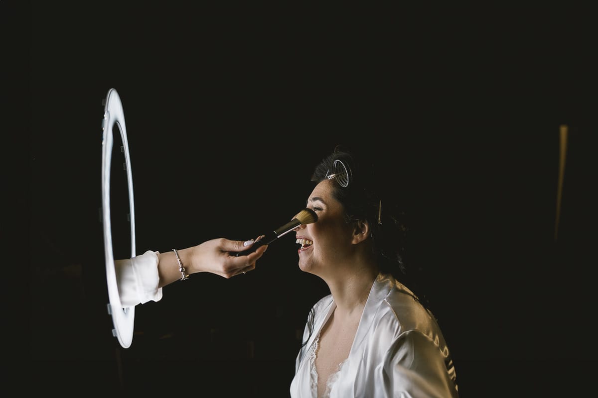 Matrimonio a Riva presso Chieri, preparazione della sposa. Make-up. Sposa che ride quando la MUA le passa un pennello sul viso attraverso una ring light