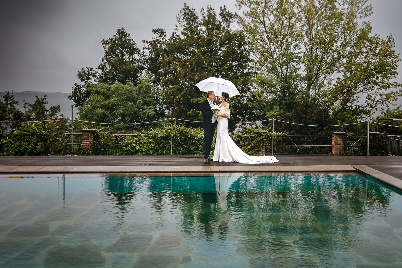 Matrimonio a "La Madernassa" nelle Langhe sotto la pioggia. Ritratti di coppia a bordo piscina sotto un ombrello bianco.