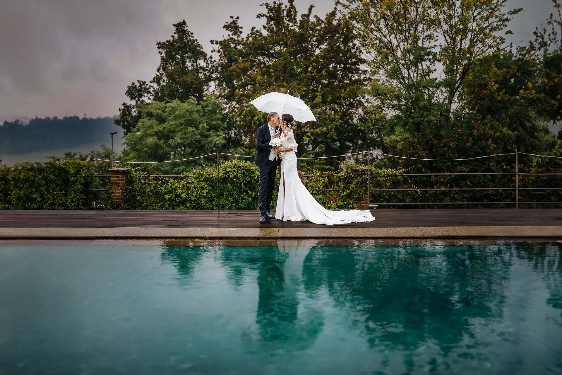Matrimonio a "La Madernassa" nelle Langhe sotto la pioggia. Ritratto degli sposi che camminano a bordo piscina sotto un ombrello bianco. Cielo gonfio di nuvole. sposi si baciano riflessi nella piscina.