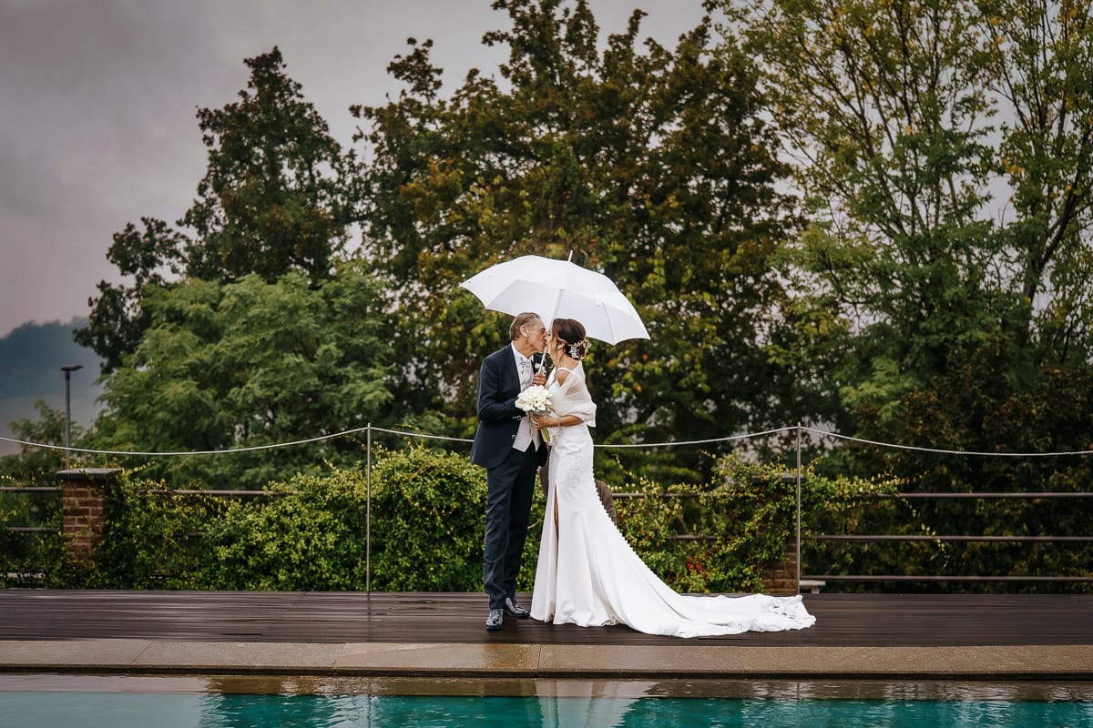 Matrimonio a "La Madernassa" nelle Langhe sotto la pioggia. Ritratti di coppia a bordo piscina sotto un ombrello bianco. Bacio.