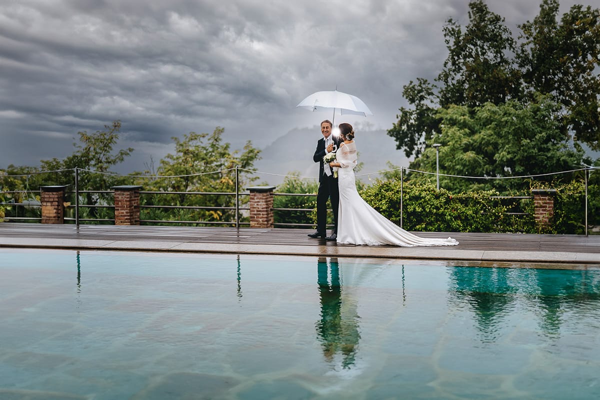 Matrimonio a "La Madernassa" nelle Langhe sotto la pioggia. Ritratti di coppia a bordo piscina sotto un ombrello bianco.