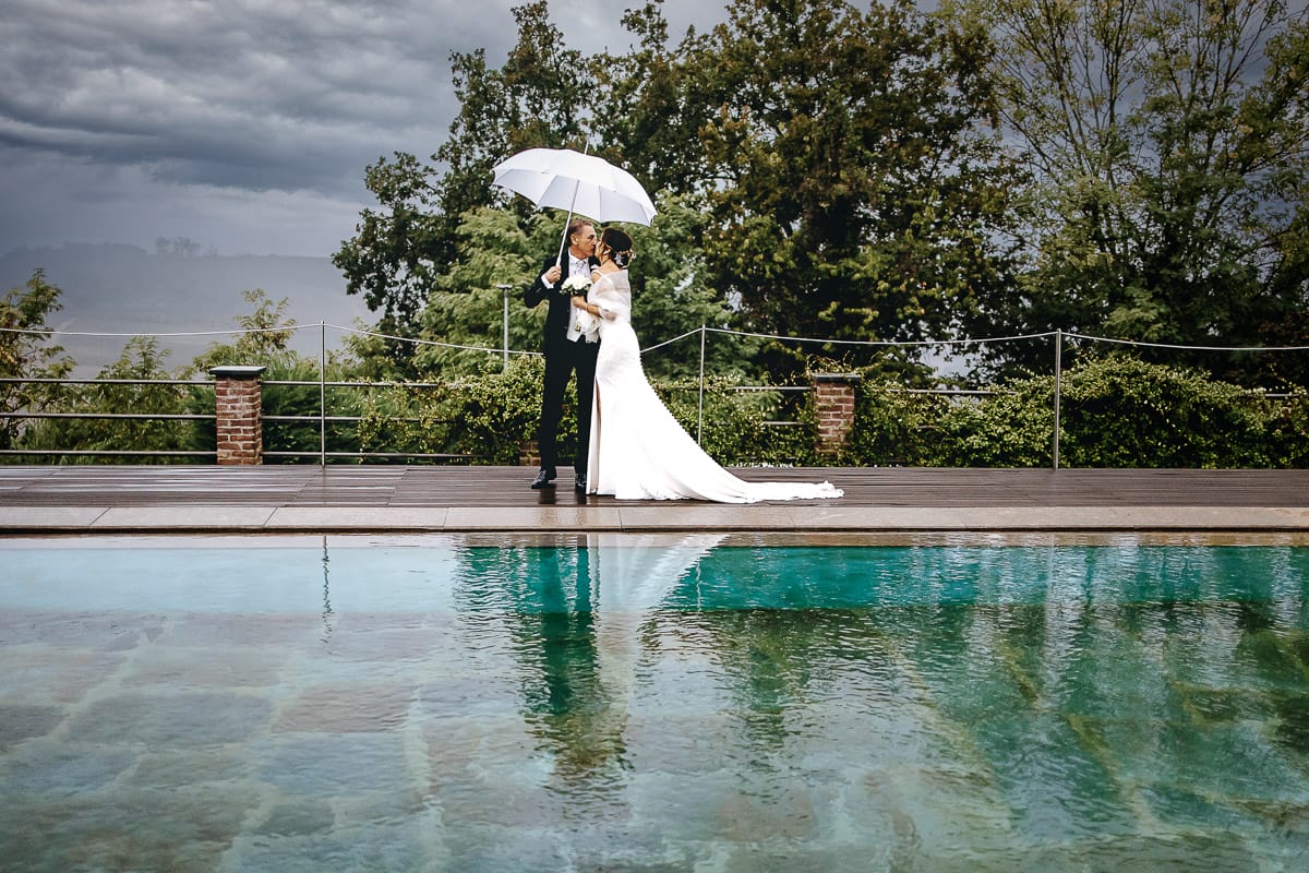 Matrimonio a "La Madernassa" nelle Langhe sotto la pioggia. Ritratti di coppia a bordo piscina sotto un ombrello bianco.