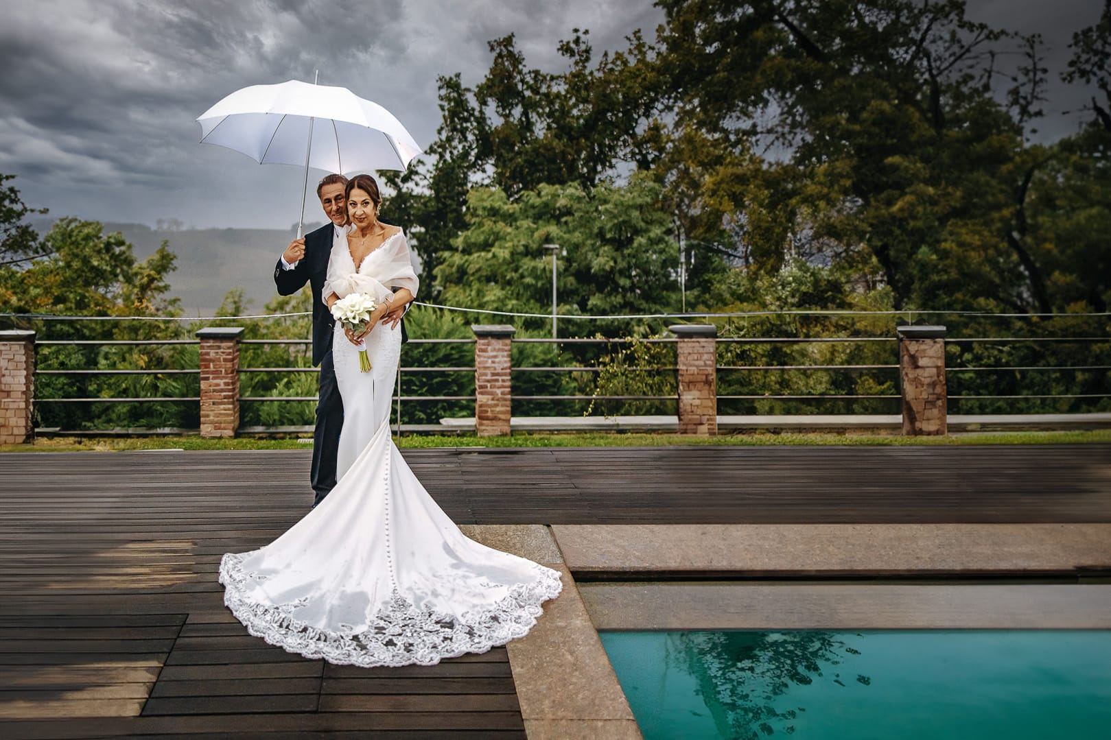 Matrimonio a "La Madernassa" nelle Langhe sotto la pioggia. Ritratti di coppia a bordo piscina sotto un ombrello bianco.