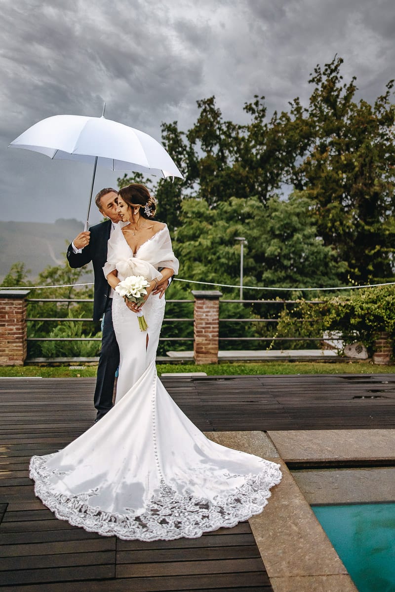 Matrimonio a "La Madernassa" nelle Langhe sotto la pioggia. Ritratti di coppia a bordo piscina sotto un ombrello bianco.