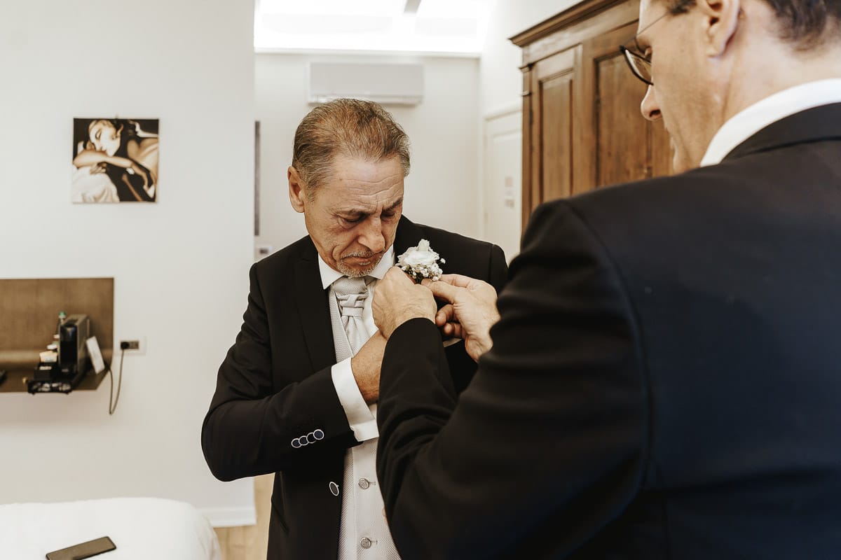 Matrimonio a "La Madernassa" nelle Langhe sotto la pioggia. preparazione dello sposo. Il testimone appunta il fiore al bavero della giacca dello sposo.