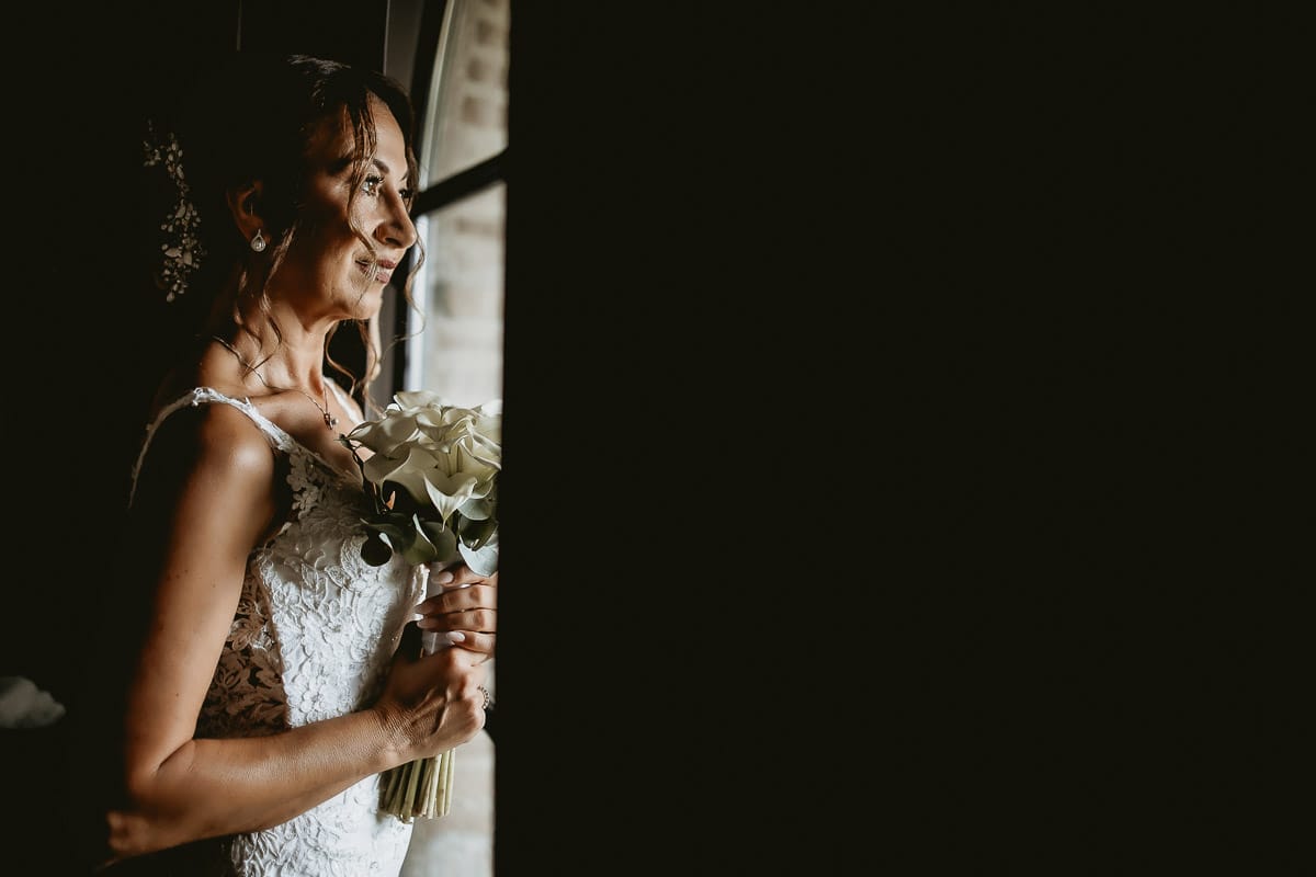 Matrimonio a "La Madernassa" nelle Langhe sotto la pioggia. Preparazione della sposa. Ritratto della sposa alla finestra con il bouquet