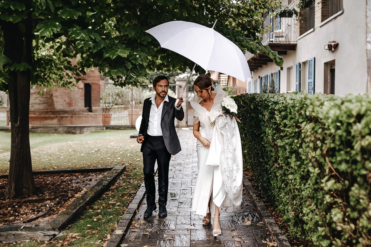 Matrimonio a "La Madernassa" nelle Langhe sotto la pioggia. Preparazione della sposa. Sposa cammina sotto l'ombrello verso l'automobile.