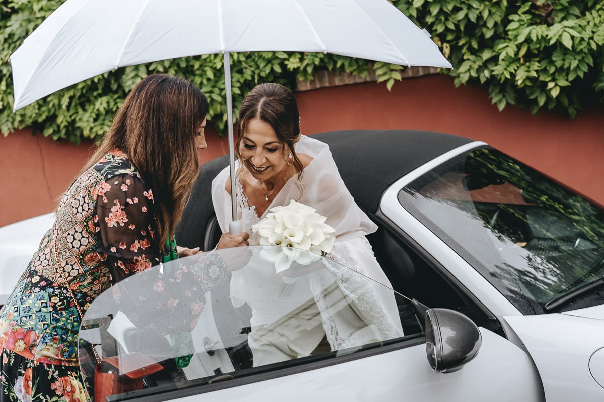 Matrimonio a "La Madernassa" nelle Langhe sotto la pioggia. Cerimonia.Sposa esce dall'auto.Wp tiene l'ombrello.