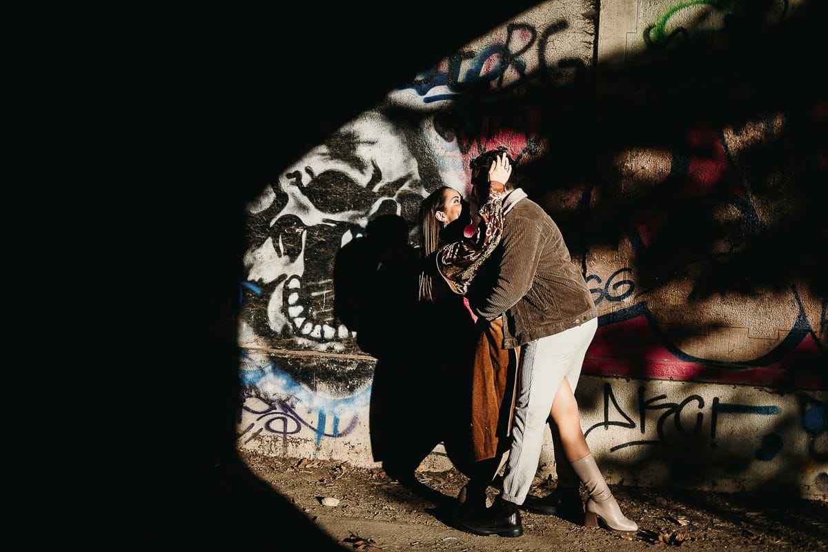 Prewedding a Torino, sul lungo Po al tramonto. Ritratto intimo della coppia sotto il ponte sul fiume. Chiaroscuro in una zona di graffiti metropolitani. Casquet