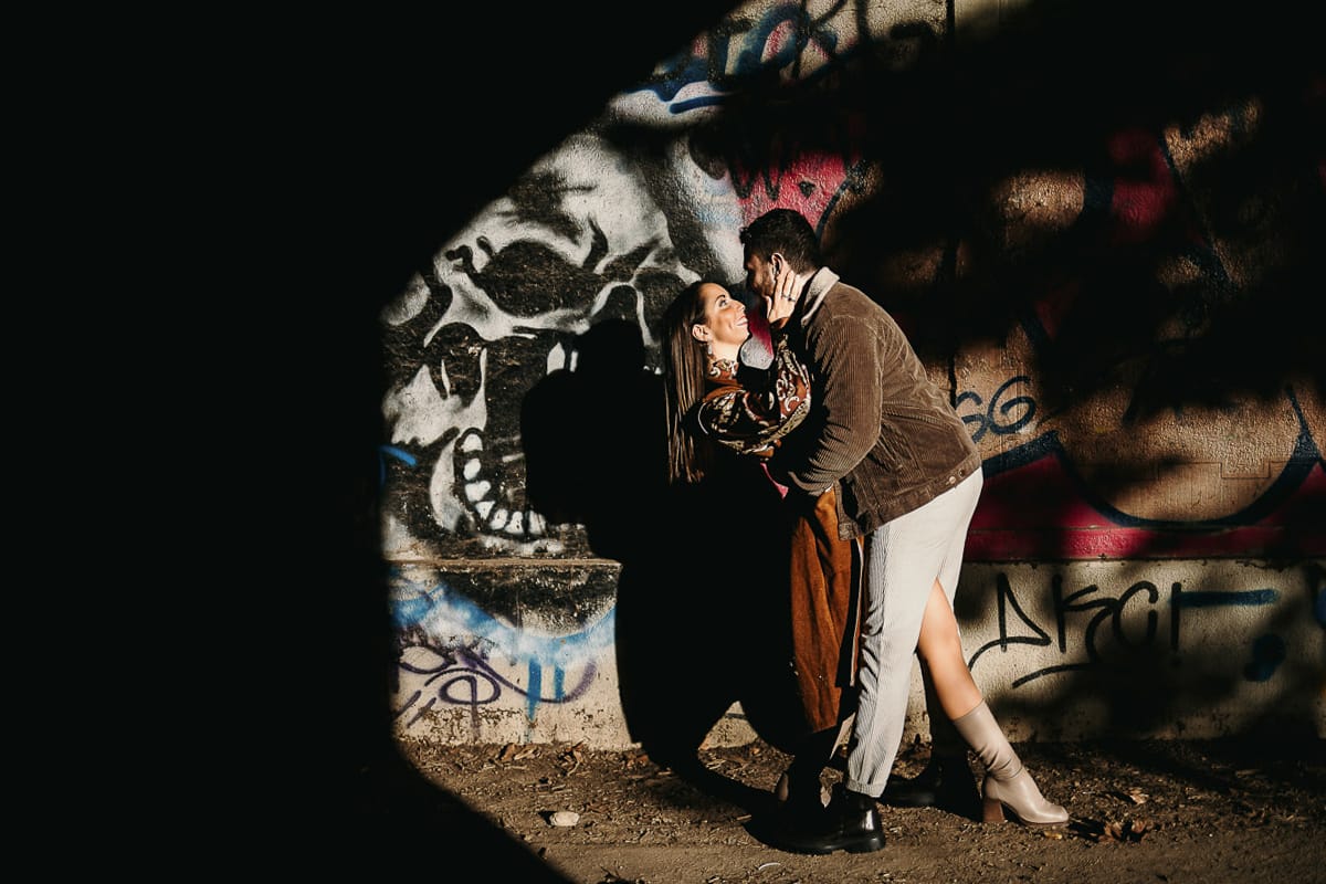 Prewedding a Torino, sul lungo Po al tramonto. Ritratto intimo della coppia sotto il ponte sul fiume. Chiaroscuro in una zona di graffiti metropolitani. Casquet.