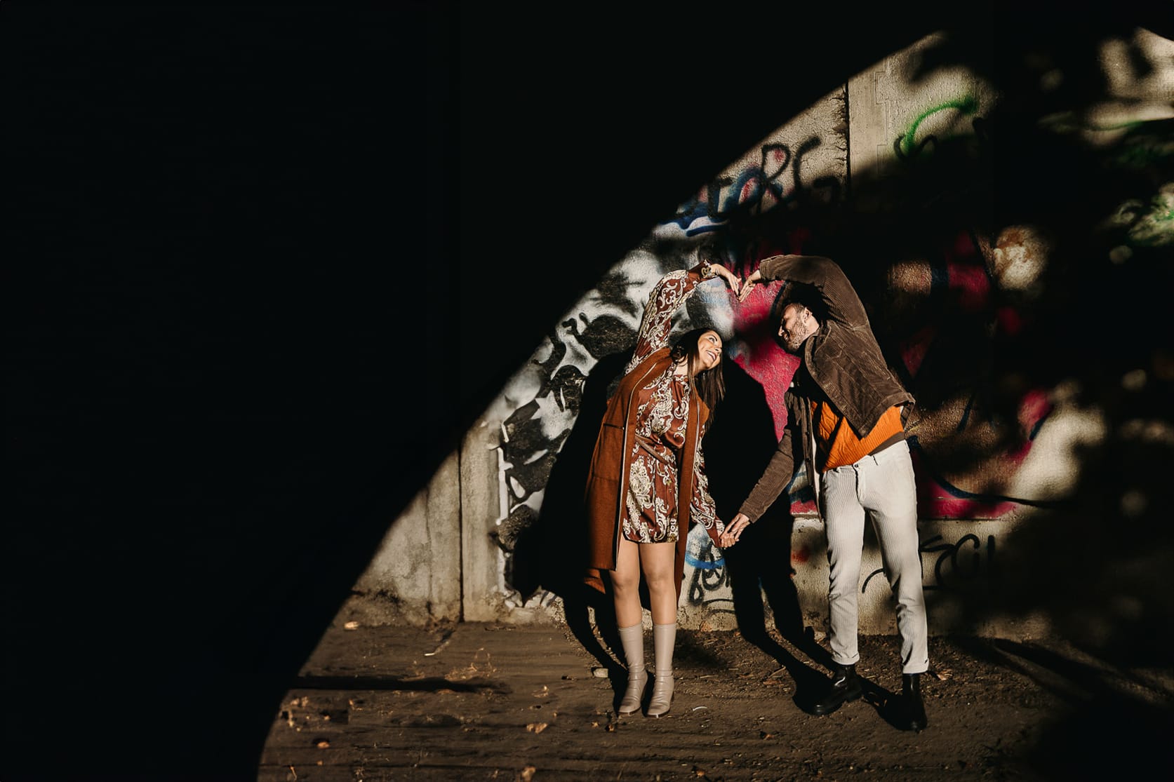 Prewedding a Torino, sul lungo Po al tramonto. Ritratto intimo della coppia sotto il ponte sul fiume. Chiaroscuro in una zona di graffiti metropolitani. la coppia crea un cuore con le braccia.