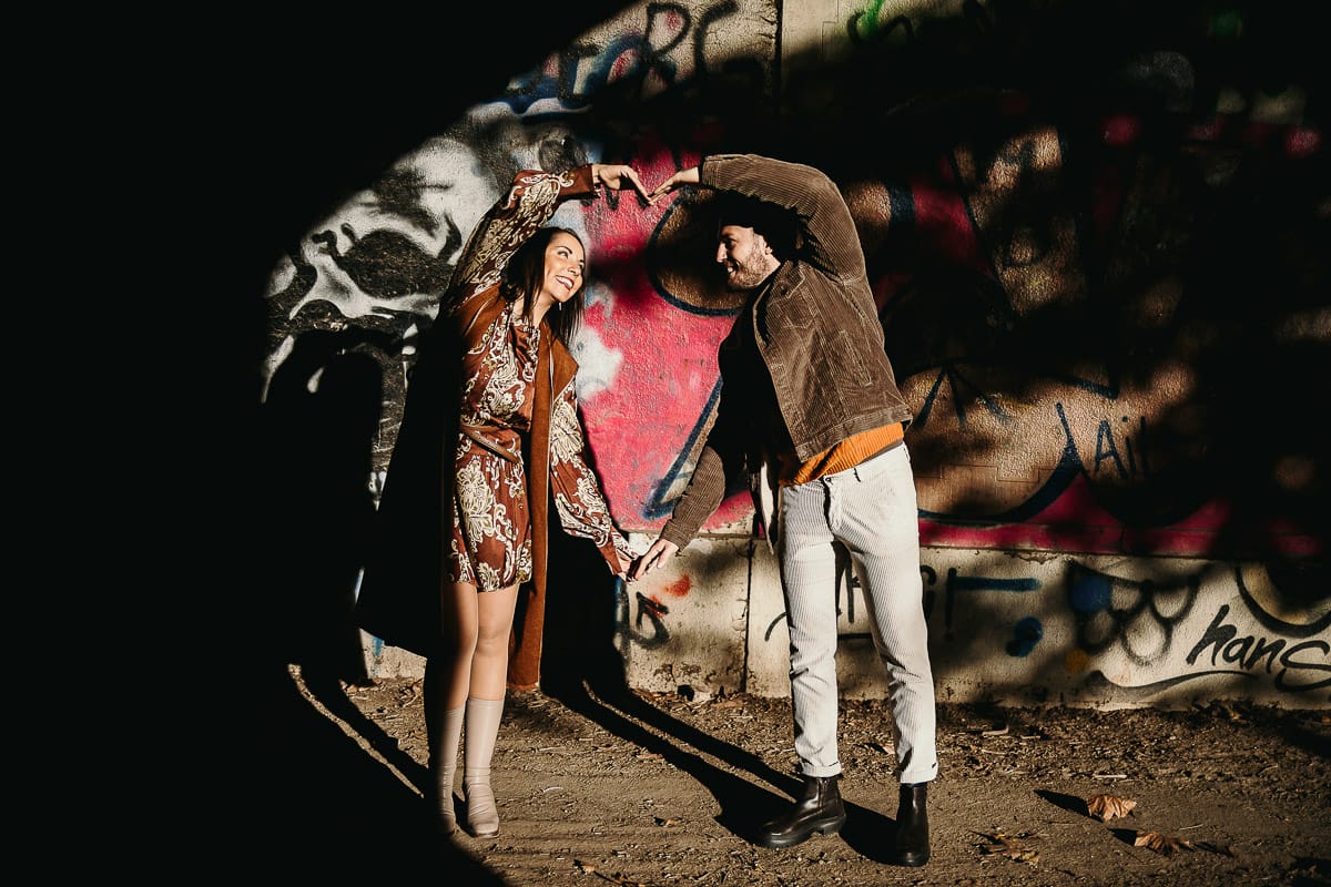 Prewedding a Torino, sul lungo Po al tramonto. Ritratto intimo della coppia sotto il ponte sul fiume. Chiaroscuro in una zona di graffiti metropolitani. la coppia crea un cuore con le braccia.