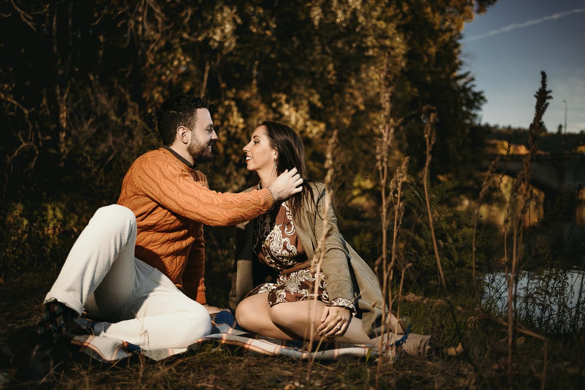 Prewedding a Torino, sul lungo Po al tramonto. Ritratto sulla riva del fiume con la magia dell'autunno sull'acqua. Coppia seduta si accarezza.