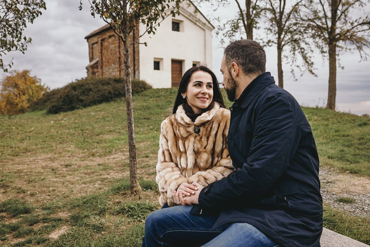 Fotografia di coppia invernale presso la chiesa di San Lorenzo di Mombello di Torino. Barbara e Riccardo seduti su di una panchina di pietra davanti alla chiesetta di san Lorenzo si guardano