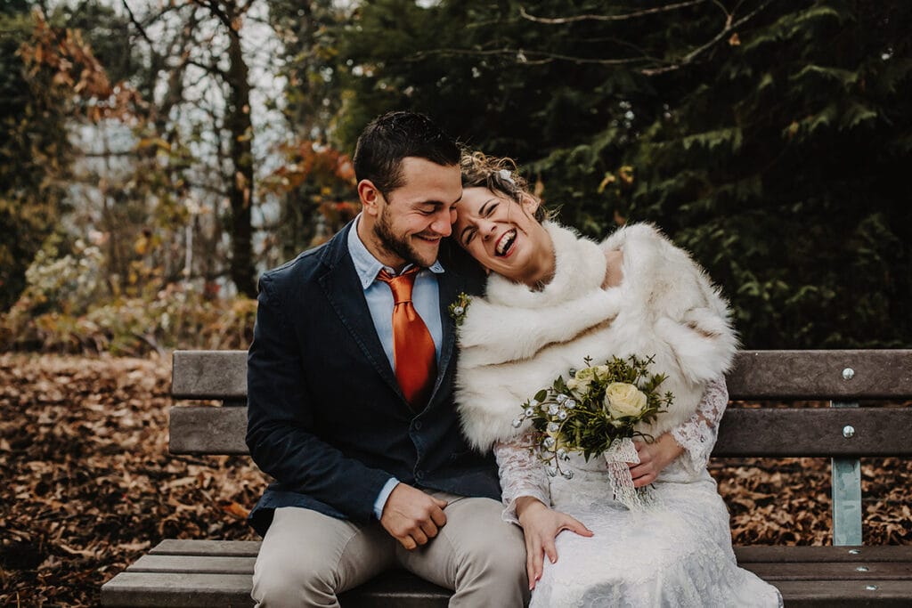 Winter wedding at the Superga hill park, among coniferous trees reminiscent of the great parks of North America. The bride and groom sitting on a wooden bench laugh happily.