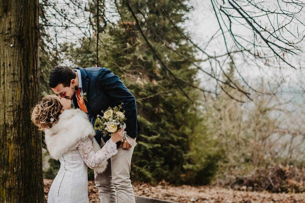 Winter wedding at the Superga hill park, among coniferous trees reminiscent of the great parks of North America. The groom standing on the bench kisses the bride.