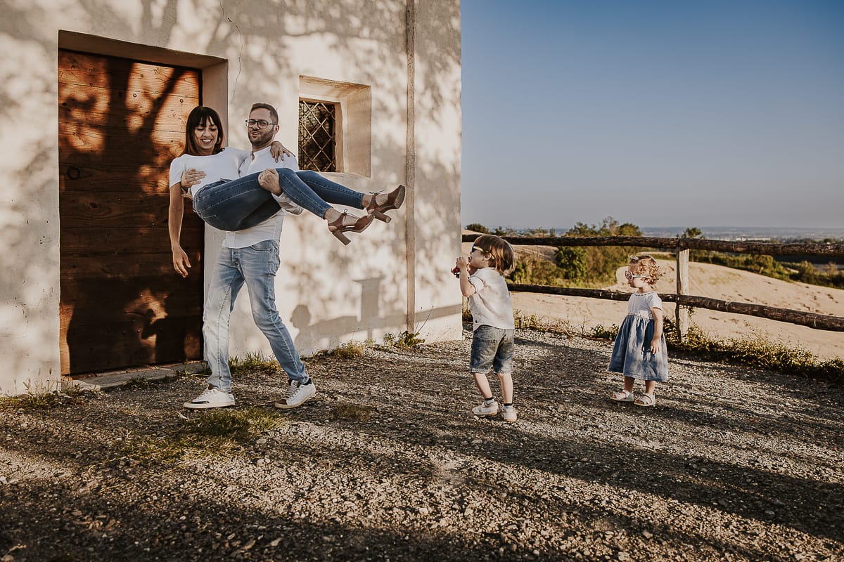 Ilenia e Filippo si stringono in un abbraccio d'amore, mentre i loro figli giocano felici al tramonto.