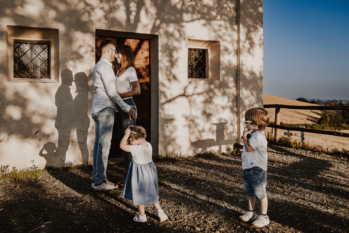 Ilenia e Filippo si stringono in un abbraccio d'amore, mentre i loro figli giocano felici al tramonto.