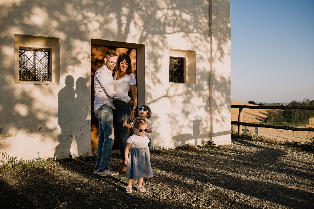 Ilenia e Filippo si stringono in un abbraccio d'amore, mentre i loro figli giocano felici al tramonto.