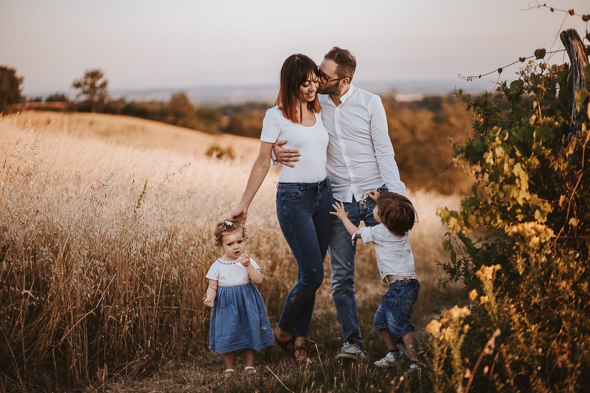 Ilenia e Filippo si stringono in un abbraccio d'amore, mentre i loro figli giocano felici al tramonto.