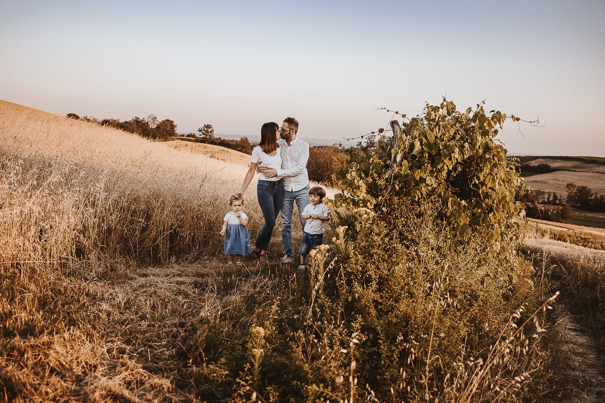 Ilenia e Filippo si stringono in un abbraccio d'amore, mentre i loro figli giocano felici al tramonto.