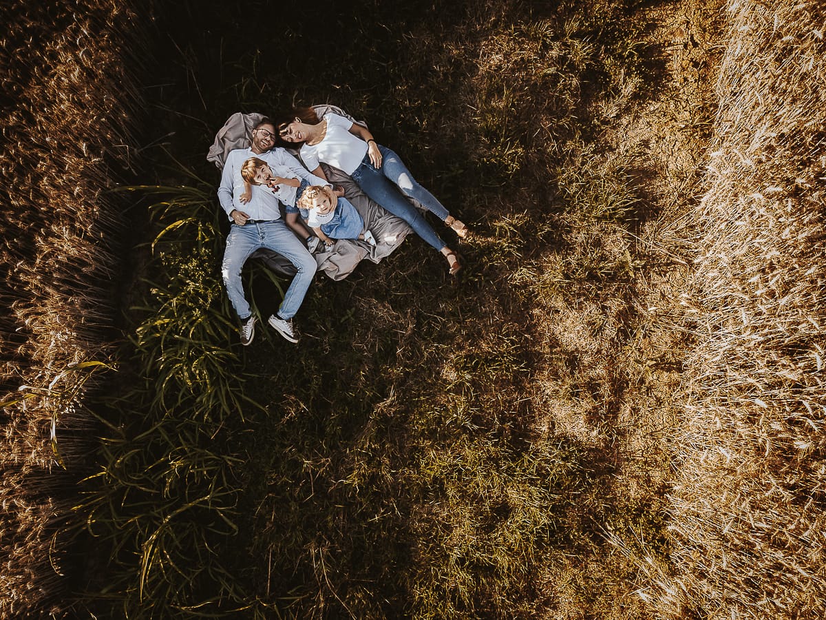 Foto di Famiglia al Tramonto: Ilenia, Filippo, Edoardo e Camilla a Mombello di Torino. la famiglia sdraiata nel sentiero in mezzo a due campi di grano. I bambini sono incuriositi dal drone.