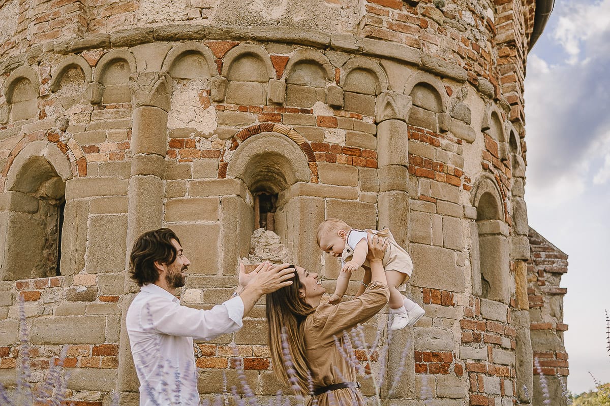 Servizio fotografico di famiglia a Torino: un tramonto dorato a Mombello di Torino. Chiara, Alessandro e leonardo in mezzo ad un cespuglio di fiori di colore lilla davanti alla chiesa romanica di San Lorenzo