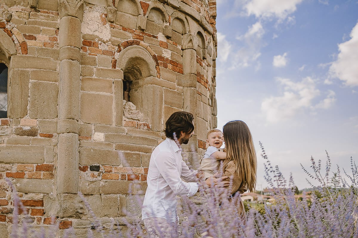 Servizio fotografico di famiglia a Torino: un tramonto dorato a Mombello di Torino. Chiara, Alessandro e leonardo in mezzo ad un cespuglio di fiori di colore lilla davanti alla chiesa romanica di San Lorenzo
