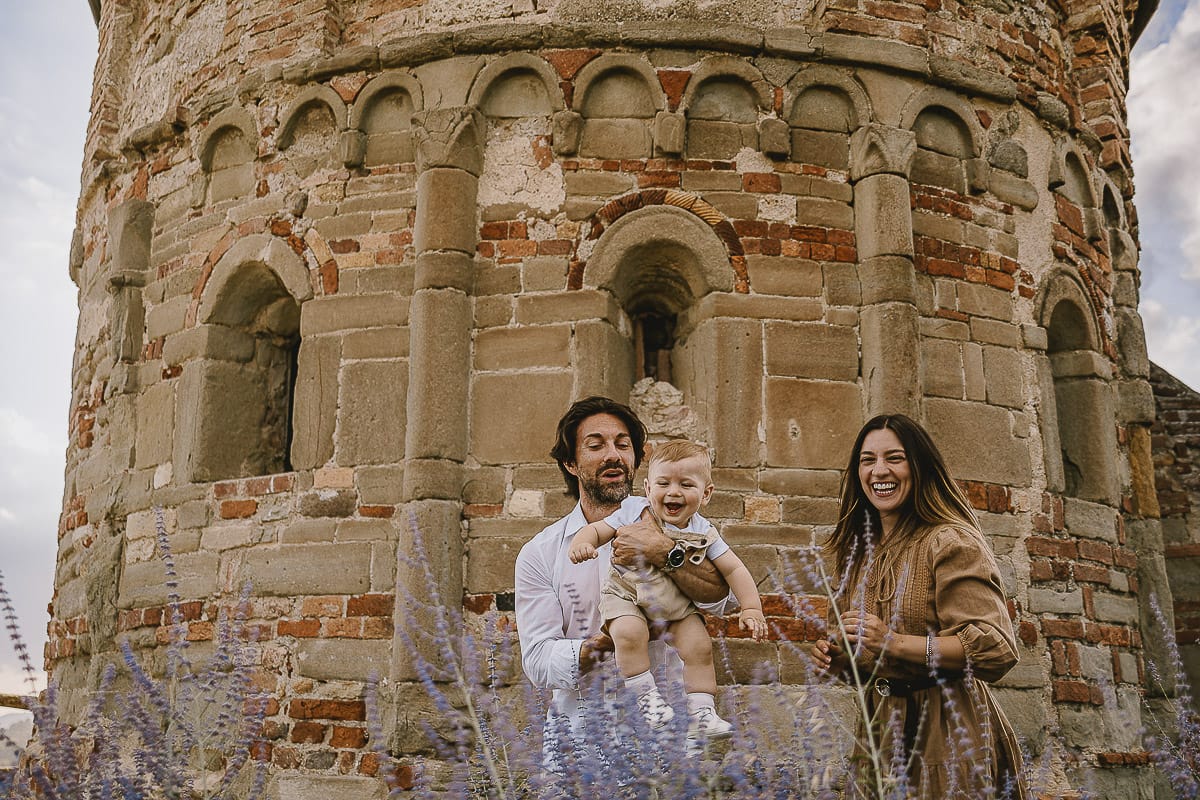 Servizio fotografico di famiglia a Torino: un tramonto dorato a Mombello di Torino. Chiara, Alessandro e leonardo in mezzo ad un cespuglio di fiori di colore lilla davanti alla chiesa romanica di San Lorenzo