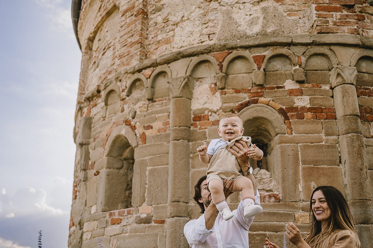 Servizio fotografico di famiglia a Torino: un tramonto dorato a Mombello di Torino. Chiara, Alessandro e leonardo in mezzo ad un cespuglio di fiori di colore lilla davanti alla chiesa romanica di San Lorenzo