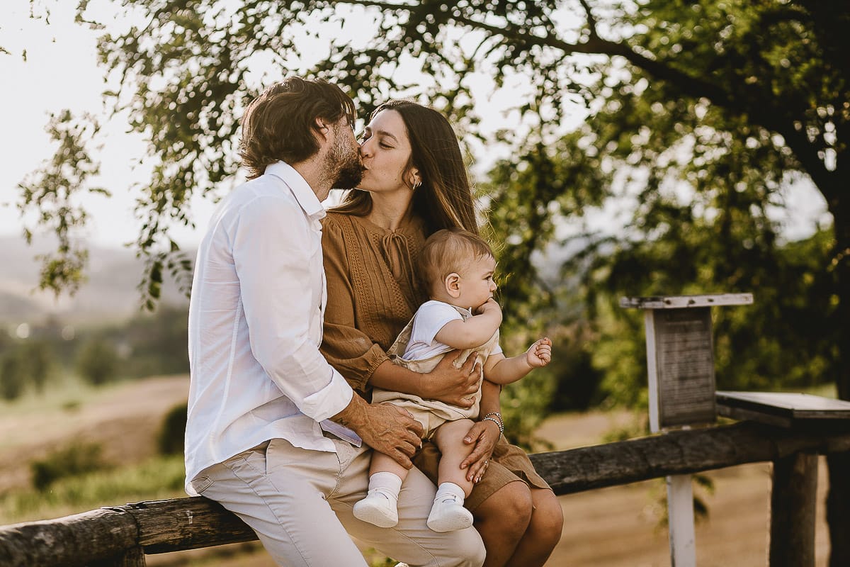 Servizio fotografico di famiglia a Torino: un tramonto dorato a Mombello di Torino. Chiara, Alessandro e il piccolo Leonardo seduti su di una staccionata. I genitori si baciano