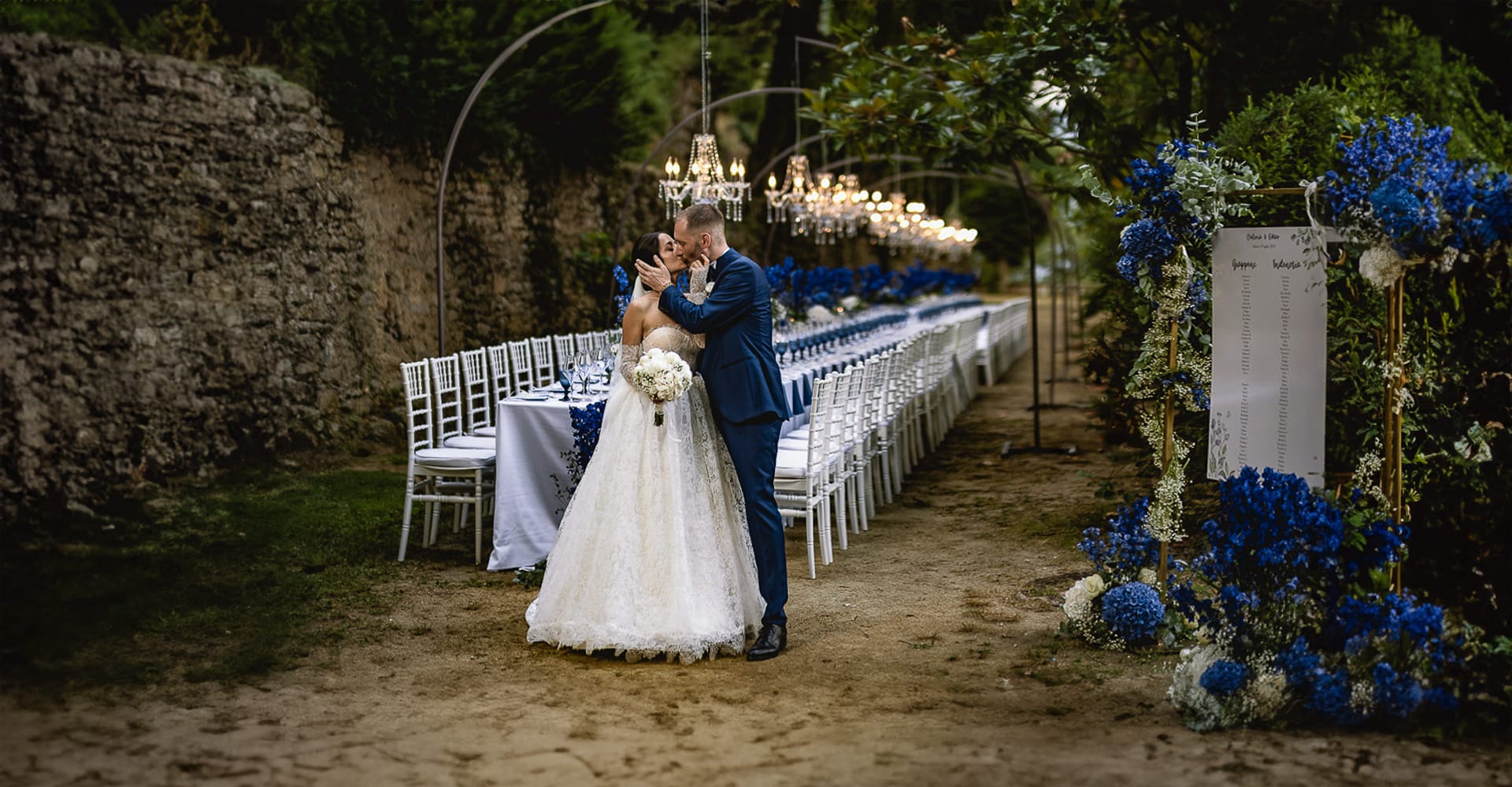 Matrimonio da favola al Castello Canali. Valeria e Omar si baciano davanti al loro bellissimo tavolo imperiale in un viale del parco. Fiori e biccheri sui toni del blu. Lampadari a goccia.