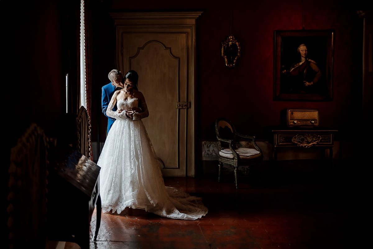 Matrimonio da favola al castello Canalis. Ritratto degli sposi nelle sale del castello. Sposi di fianco alla porta finestra. Sposo bacia la sposa sul collo.