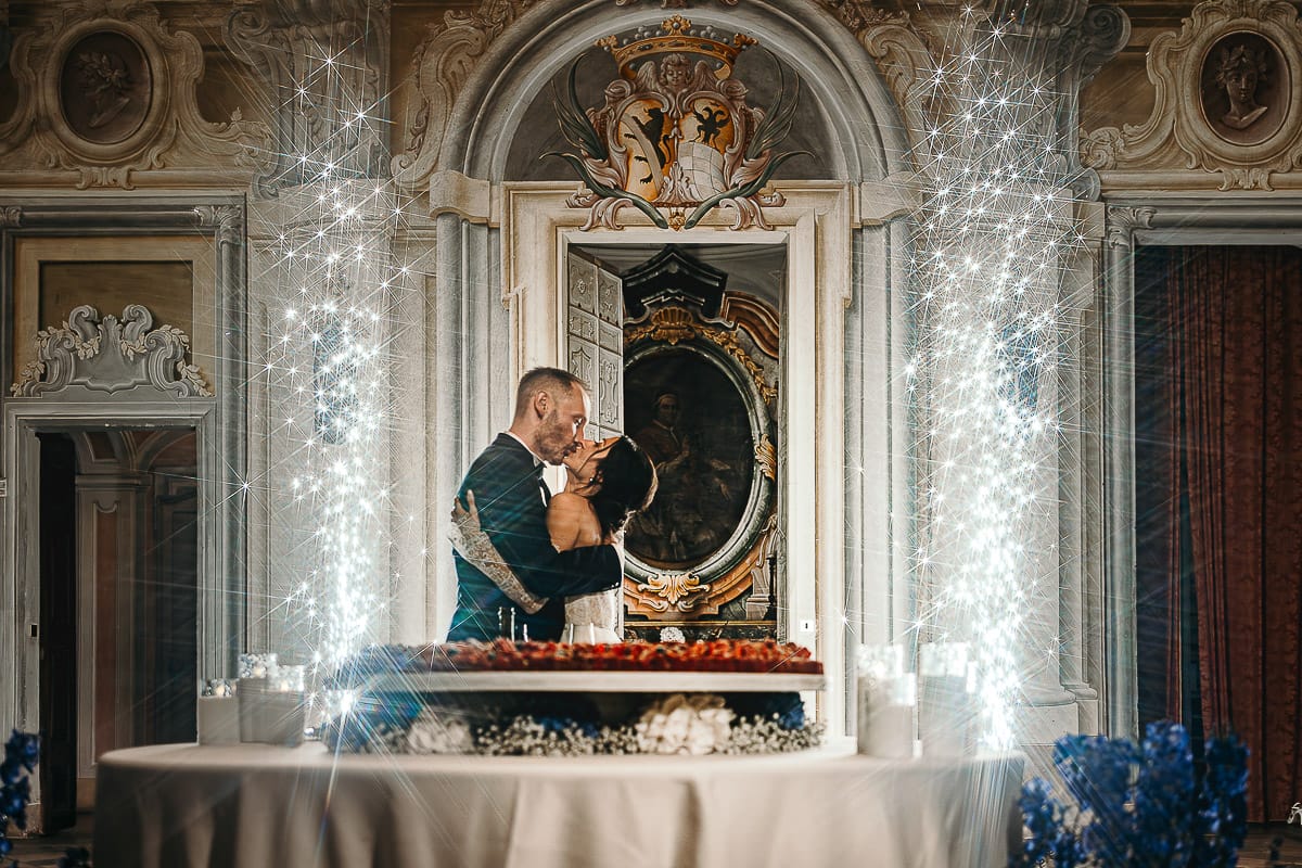 Matrimonio da favola al castello Canalis. Ricevimento. Taglio della torta. sposi si baciano nel salone delle feste, affrescato, dietro la crostata nuziale di Fabrizio Racca. Fontane luminose.
