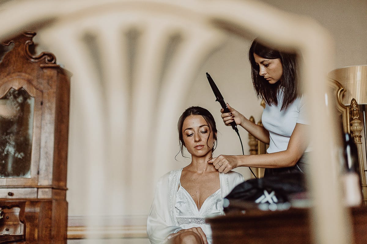 Matrimonio da favola al castello Canalis. Preparazione della sposa. La parrucchiera usa la piastra per capelli. Ritratto della sposa ripresa attraverso lo schienale di una sedia.