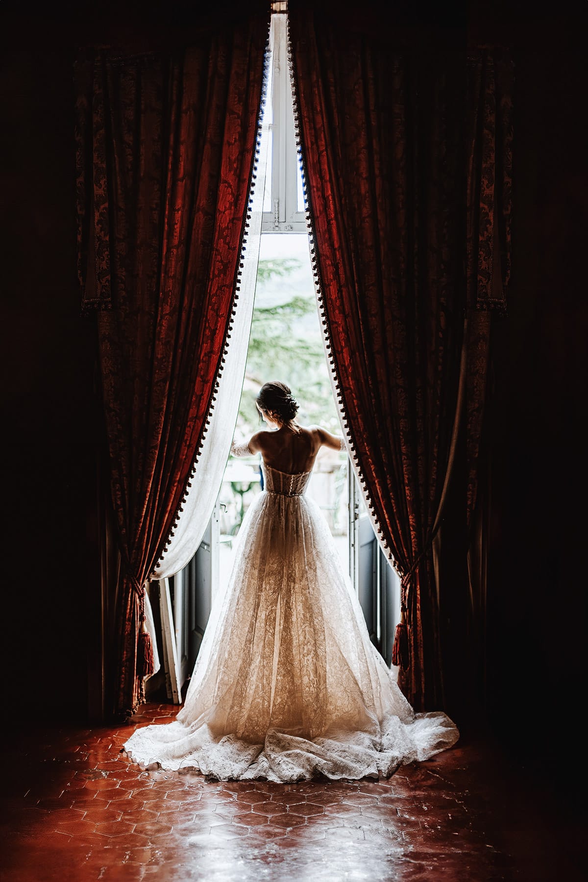 Matrimonio da favola al castello Canalis. Ritratto della sposa nella sala rossa del castello. Silhouette della sposa di fronte alla porta finestra in mezzo a due tende di velluto rosso.