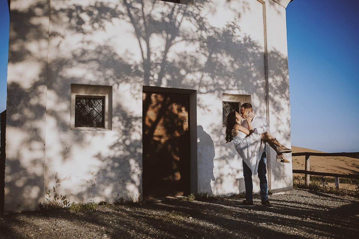 Prewedding romantico e da sogno al tramonto in Monferrato. Valeria e Omar davanti alla chiesetta di san Lorenzo. L'ombra degli alberi crea una trama d'ombra molto interessante. Omar prende Valeria in braccio.