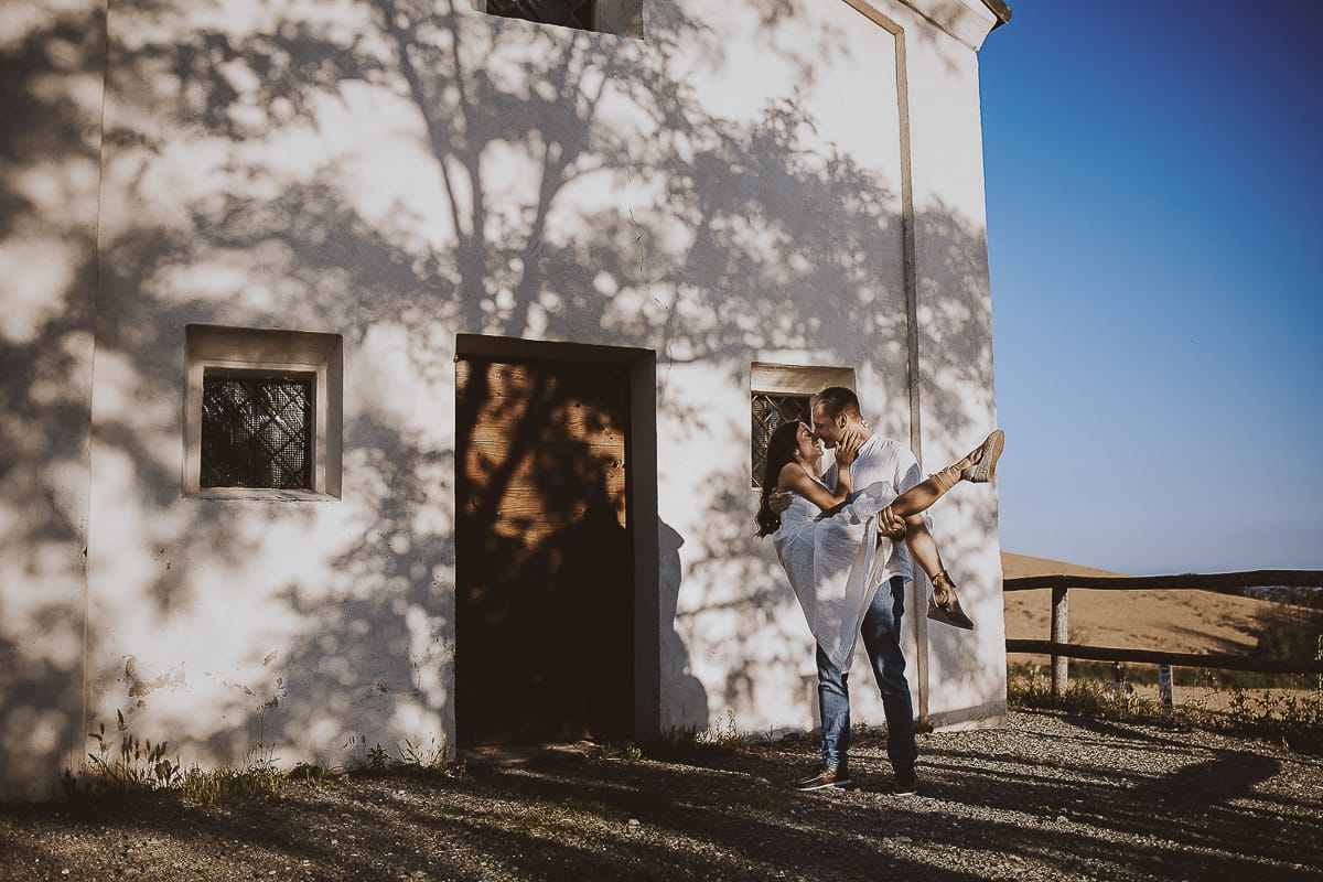 Prewedding romantico e da sogno al tramonto in Monferrato. Valeria e Omar davanti alla chiesetta di san Lorenzo. L'ombra degli alberi crea una trama d'ombra molto interessante. Omar prende Valeria in braccio.