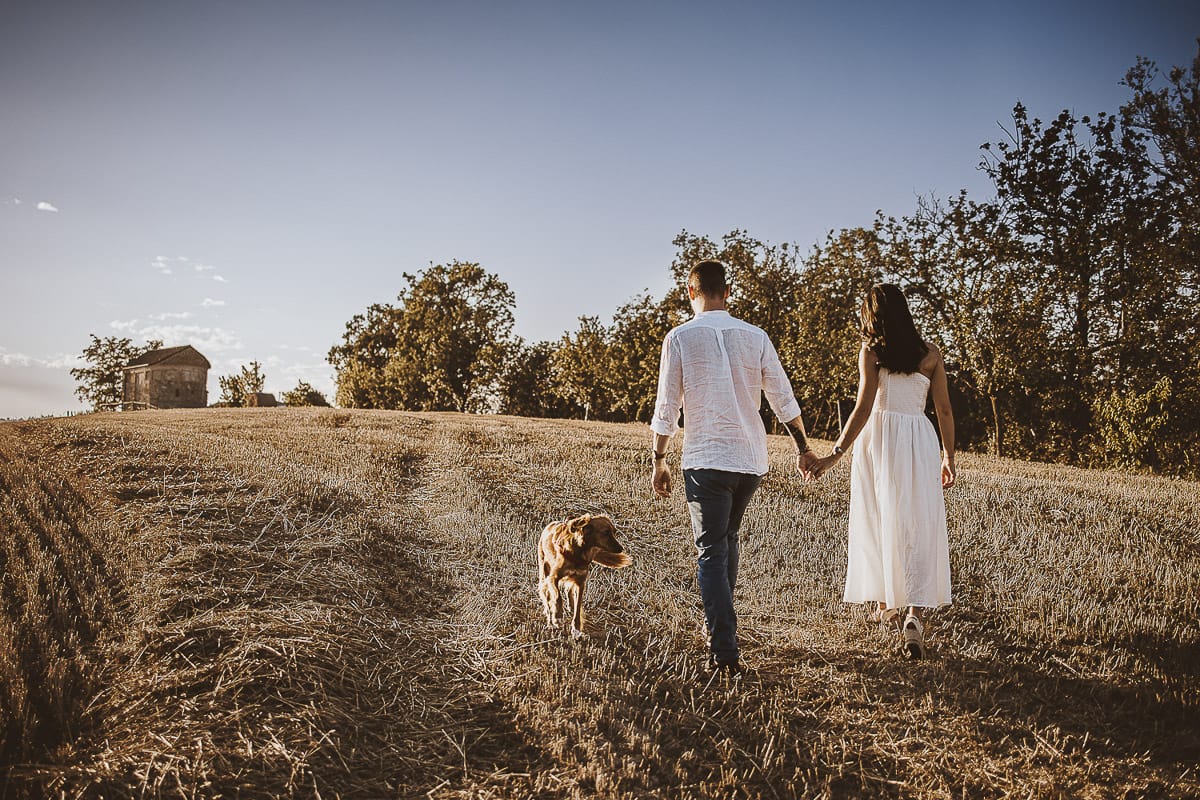 Prewedding romantico e da sogno al tramonto in Monferrato. Valeria e Omar camminano tenendosi per mano in un campo di grano tagliato. La cagnolina Nadine è davanti a loro.