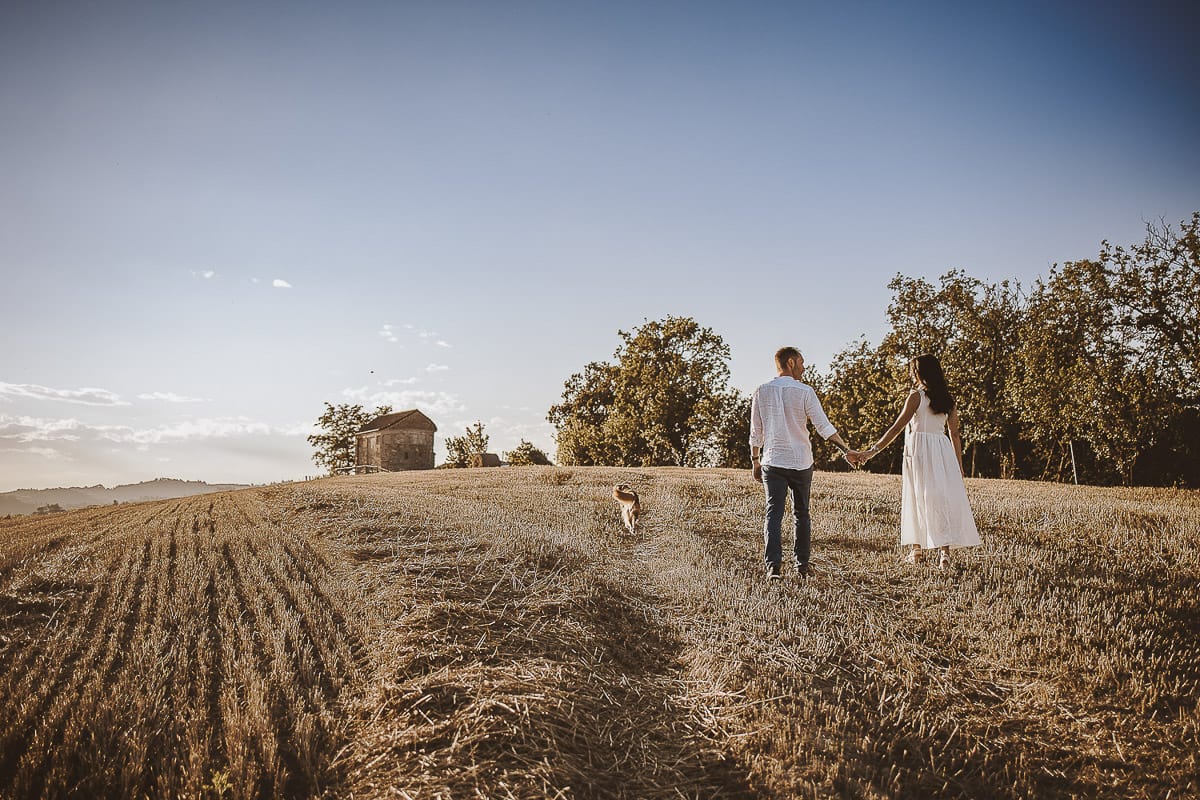 Prewedding romantico e da sogno al tramonto in Monferrato. Valeria e Omar camminano tenendosi per mano in un campo di grano tagliato.