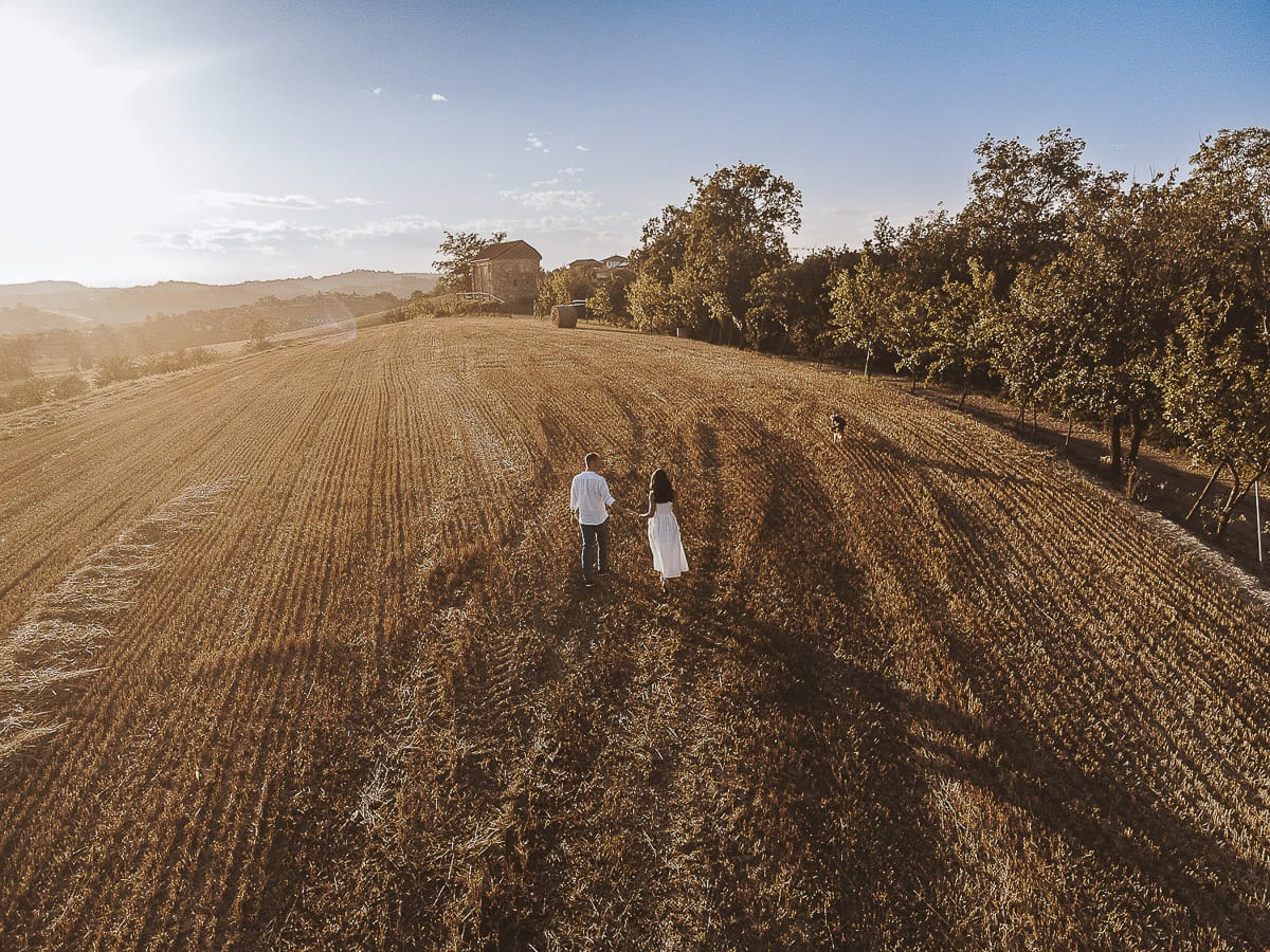Prewedding romantico e da sogno al tramonto in Monferrato. Valeria e Omar camminano tenendosi per mano in un campo di grano tagliato. Foto aerea ripresa dal drone.
