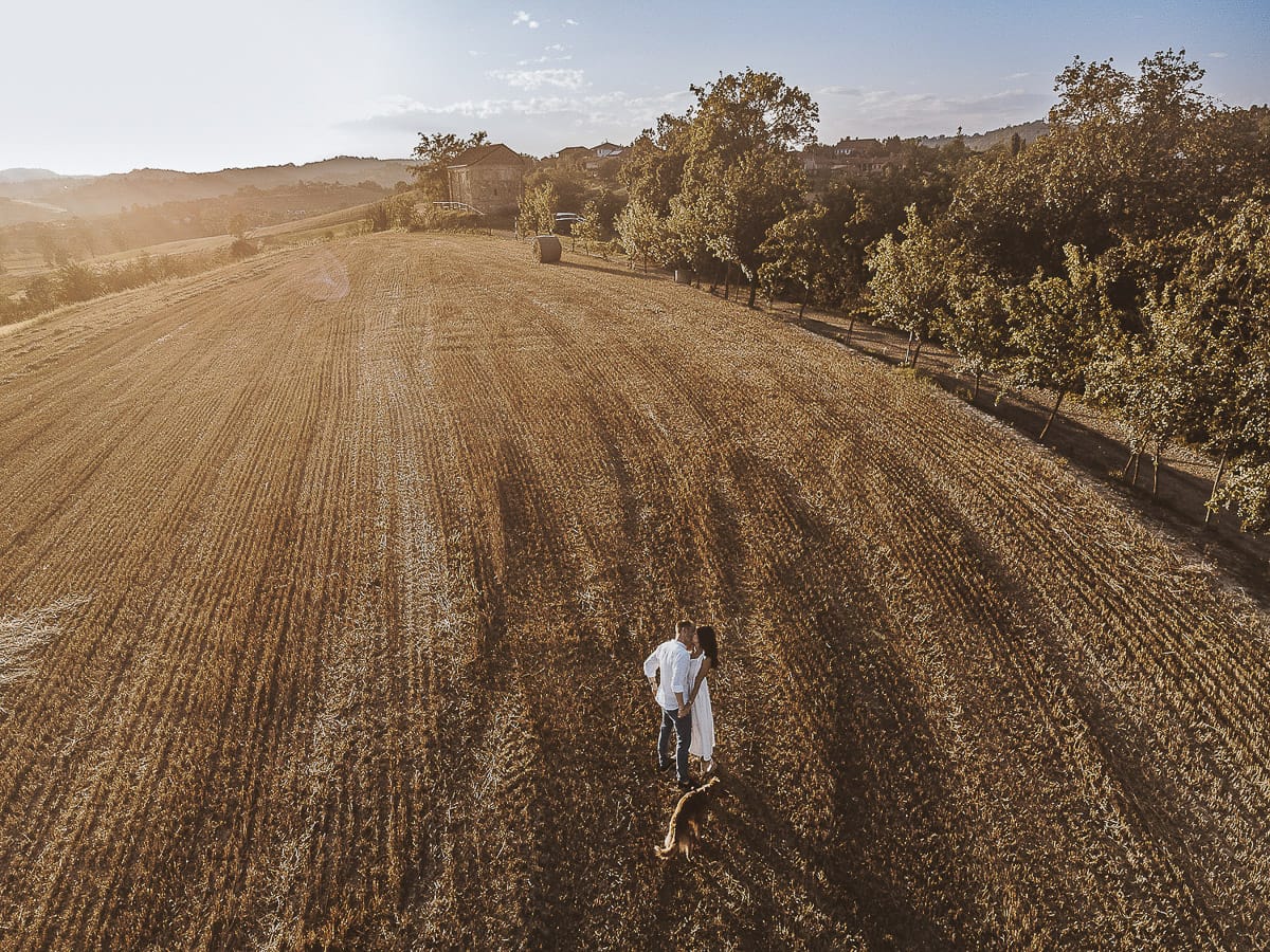 Prewedding romantico e da sogno al tramonto in Monferrato. Valeria e Omar camminano tenendosi per mano in un campo di grano tagliato. Foto aerea ripresa dal drone.